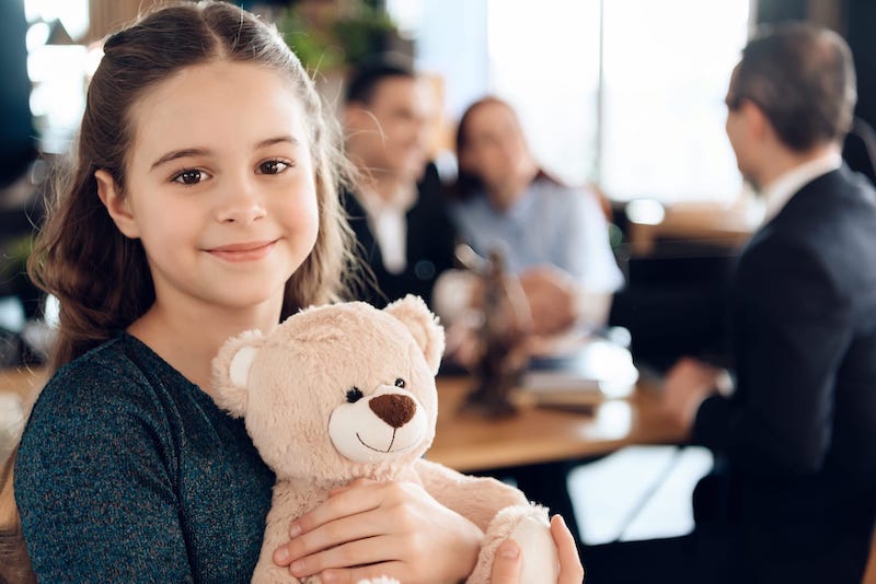 Smiling Child Holding A Stuffed Bear With Parents In The Background