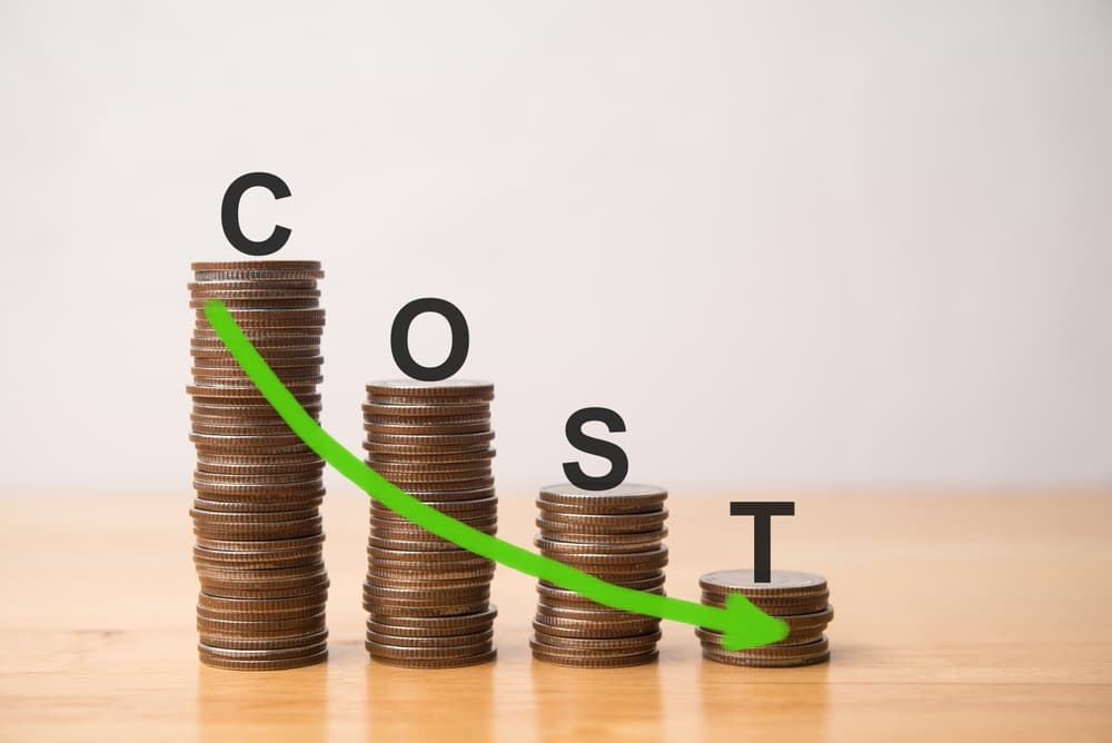 COST word and moving down arrow on decrease graph stack coins on wooden table with white wall background.