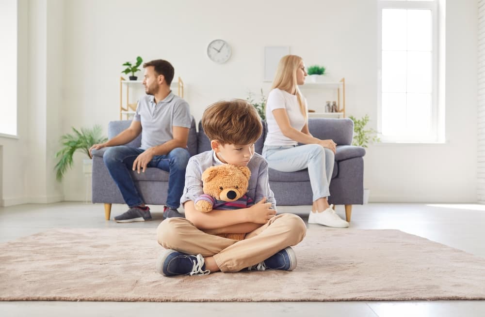 Frustrated upset boy son sitting on floor with toy bear while his mother and father sitting on opposite sides of sofa