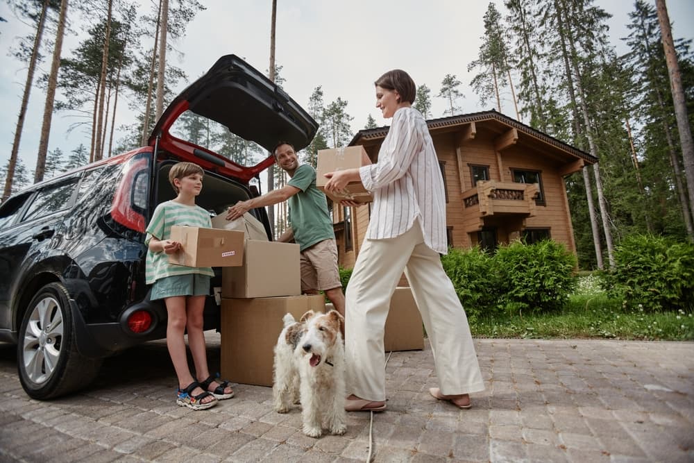 A family unloading boxes from car trunk while moving into new state with pet dog.